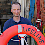 Foto von Axel Steier. Er steht hinter einem Rettungsring auf dem Lifeline steht. Im Hintergrund hängen aufgeschossene Leinen in verschiedenen Farben. 