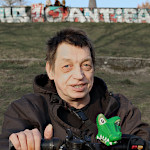 Marcus Erpenbach in seinem Rollstuhl mit Krokodil am Lenkrad im Alaunpark. Im Hintergrund eine Mauer, auf der in weißer Schrift Antifa steht. Dahinter ein Kirchturm.